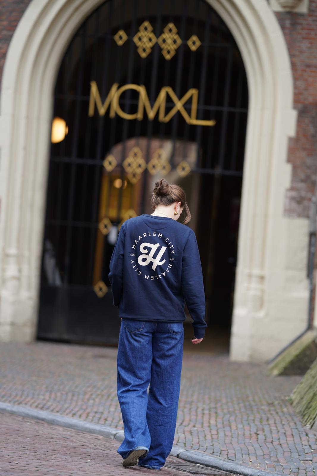 Vrouw in een donkerblauwe HIZ-sweater met Haarlem City-print op de rug, bij een historische poort in Haarlem.