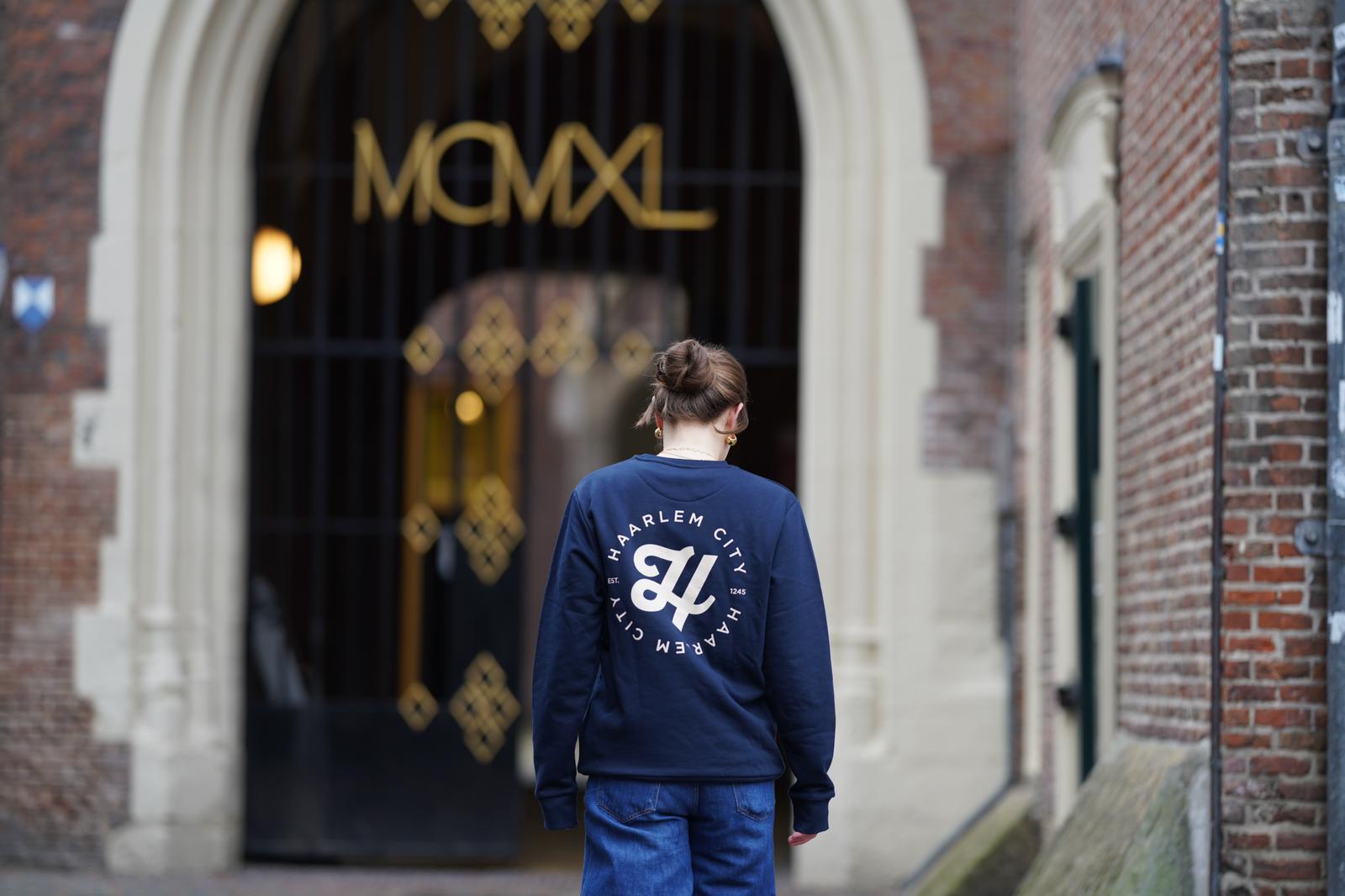 Vrouw in een donkerblauwe HIZ-sweater met Haarlem City-print op de rug, bij een historische poort in Haarlem.